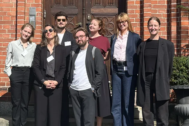 Sju personer står i solen på en trappa framför en pampig träport.