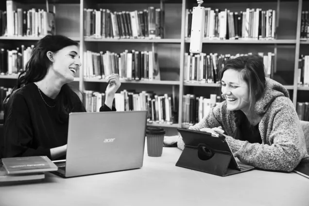 Två kvinnor på bibliotek skrattar tillsammans över sina laptops. Foto.
