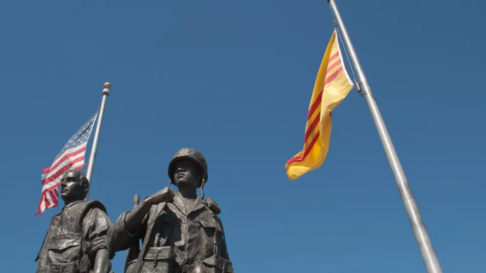 Photo of a statue of two soldiers next to the American and South Vietnamese flags against a blue sky.
