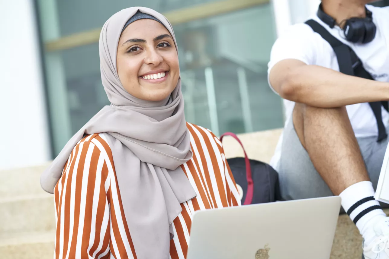 Woman in grey headscarf and orange stripy shirt smiles behind laptop. Photo. 