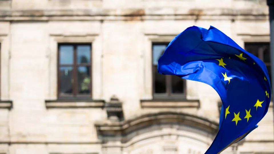 EU-flagga blåser i vinden framför byggnad. Foto: Markus Spiske, unsplash.