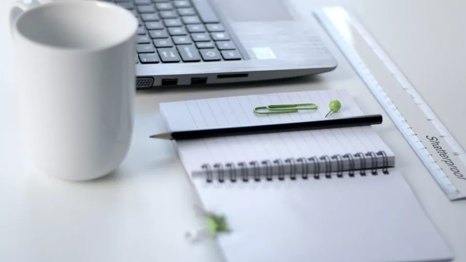 Bild på skrivbord med laptop, penna, block, kaffekopp och linjal.