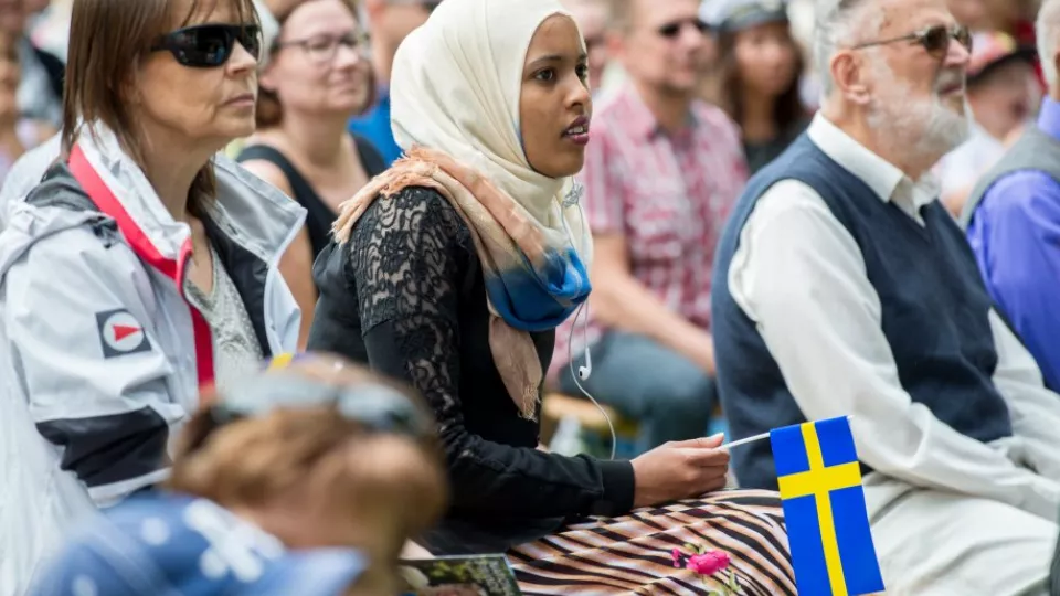 Mångfald av människor varav ena håller i svenska flaggan. 