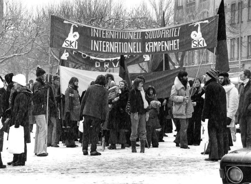Ett svartvitt fotografi på personer som står ute i snö och håller en banderroll som proklamerar internationell soldaritet.